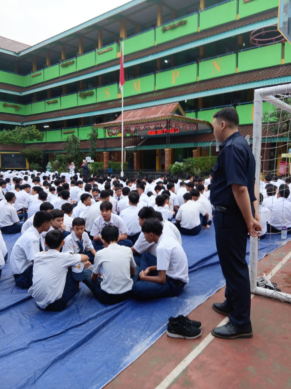 Pendamping berdiri di sisi lapangan saat murid berkumpul di atas alas biru.