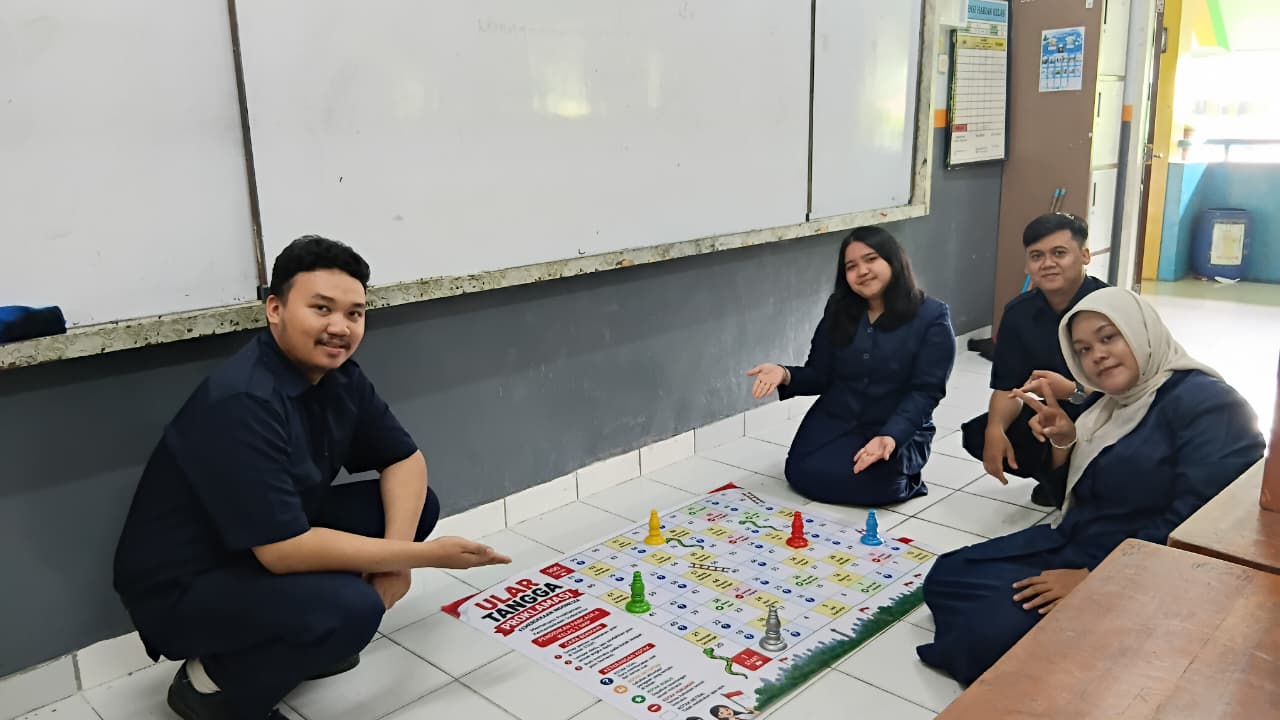 Empat orang berfoto bersama papan permainan ular tangga edukatif di kelas.